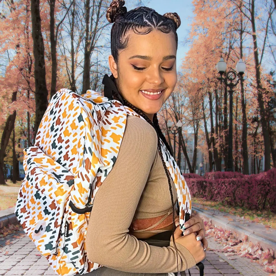 A woman wearing a tan ribbed long-sleeve top and black braids, smiling while wearing a large patterned baby carrier backpack on her shoulders in an outdoor park setting with autumn trees.