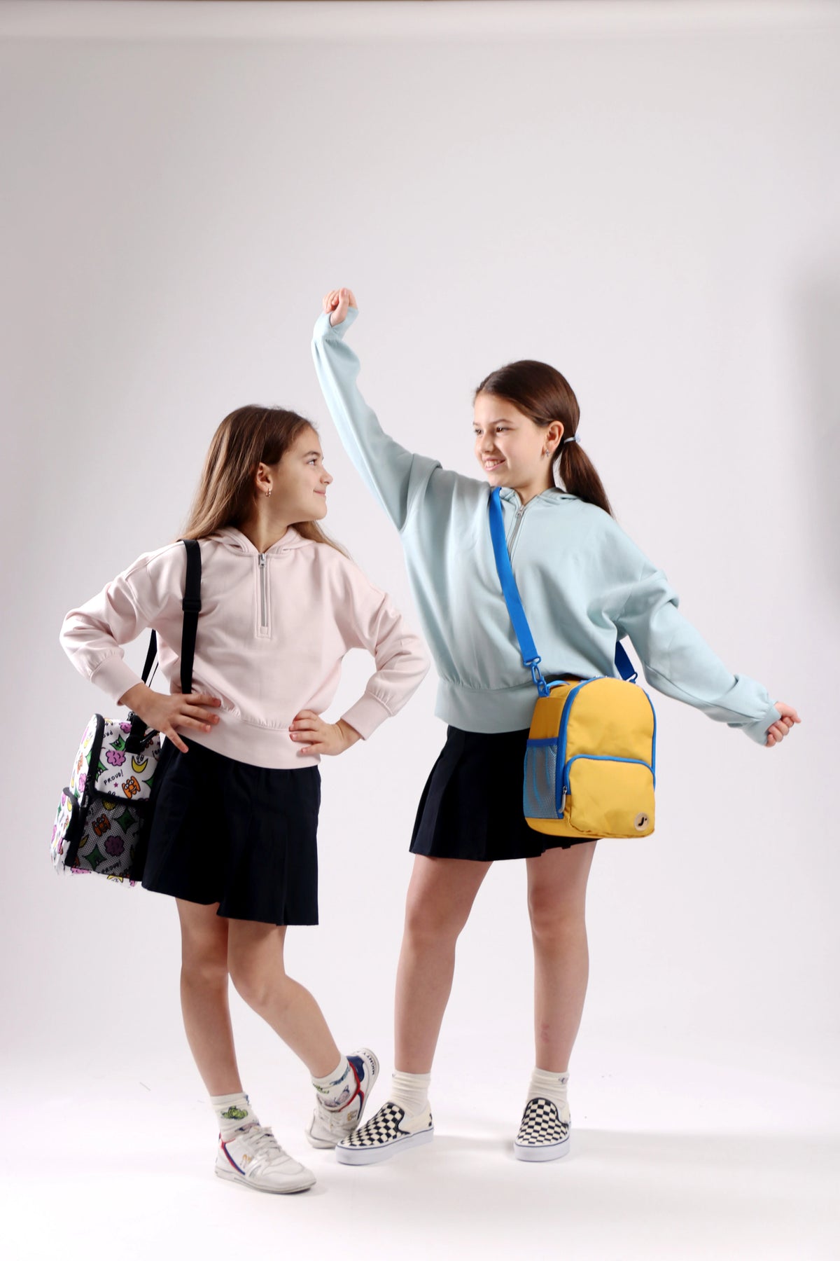Two young girls modeling school bags in a studio setting. The girl on the left wears a light pink hoodie and black skirt, carrying a black patterned shoulder bag.