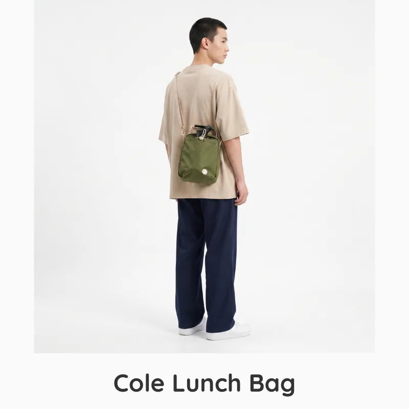 A model shown from the back wearing a beige t-shirt, navy pants, and white sneakers, carrying an olive green Cole Lunch Bag crossbody bag. The image is set against a plain white studio background with the text 'Cole Lunch Bag' printed at the bottom.