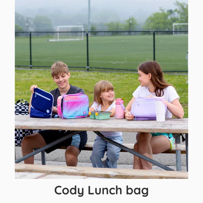 Children sitting at a picnic table with colorful lunch bags and bottles, with a grassy field and fence in the background.