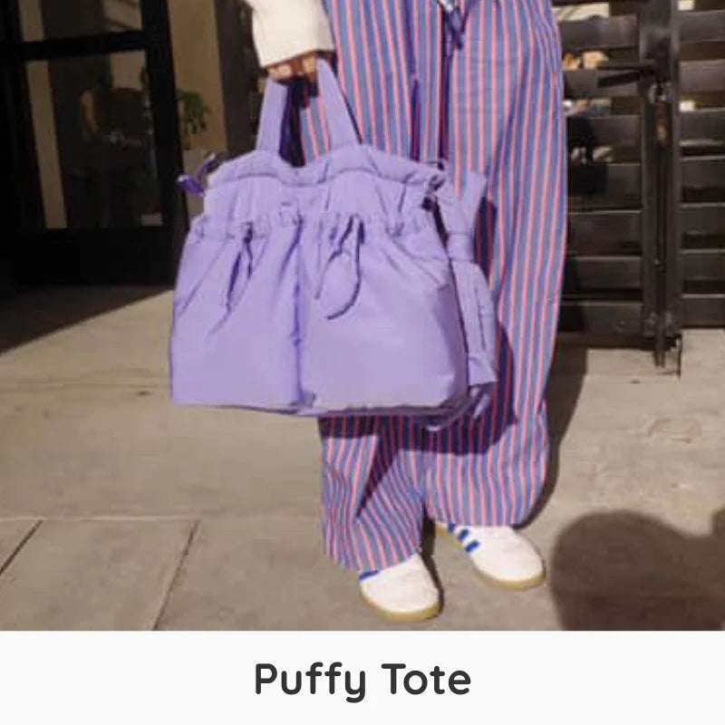 A person wearing loose pink, blue, and white striped pants holds a lavender purple puffy drawstring tote bag outdoors on a sidewalk. The text 'Puffy Tote' appears at the bottom.